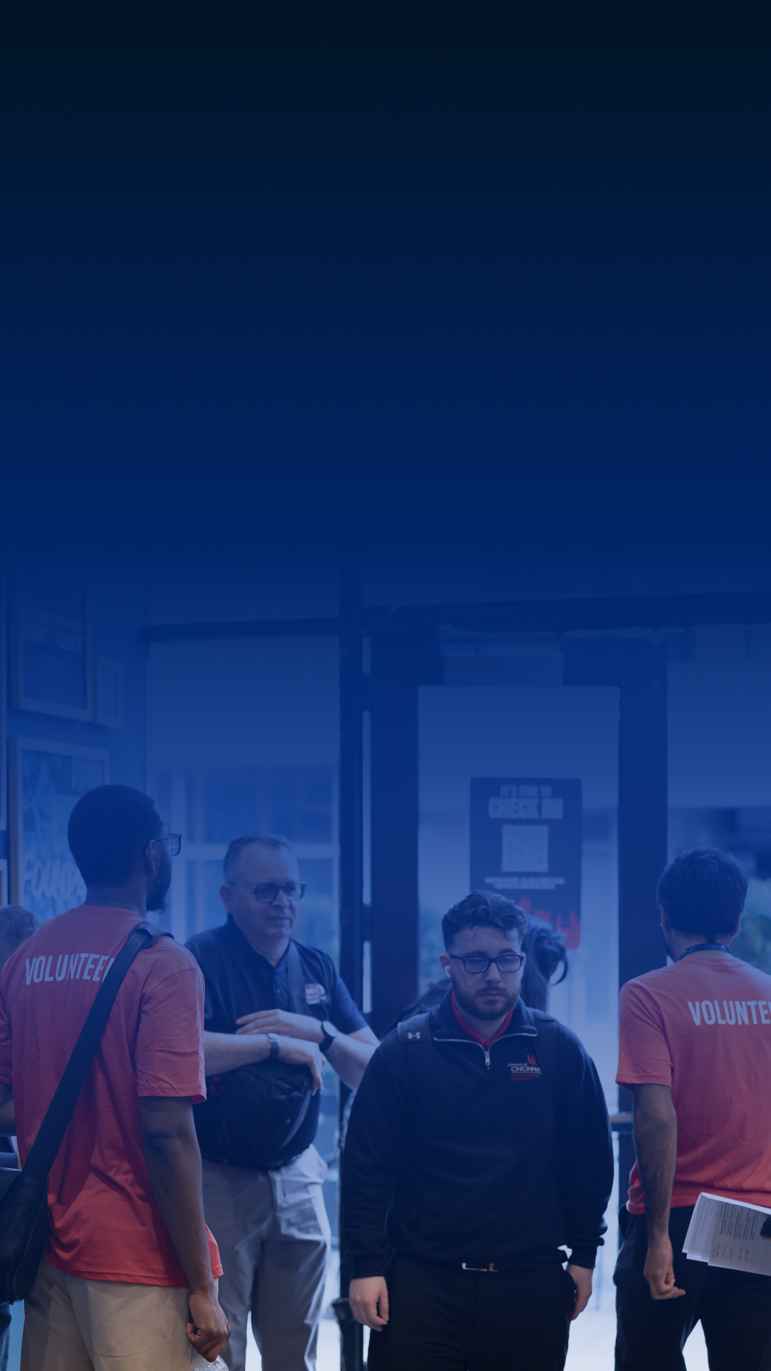 Group of people standing inside a building, some wearing 'Volunteer' shirts, with a glass door and informational posters in the background.