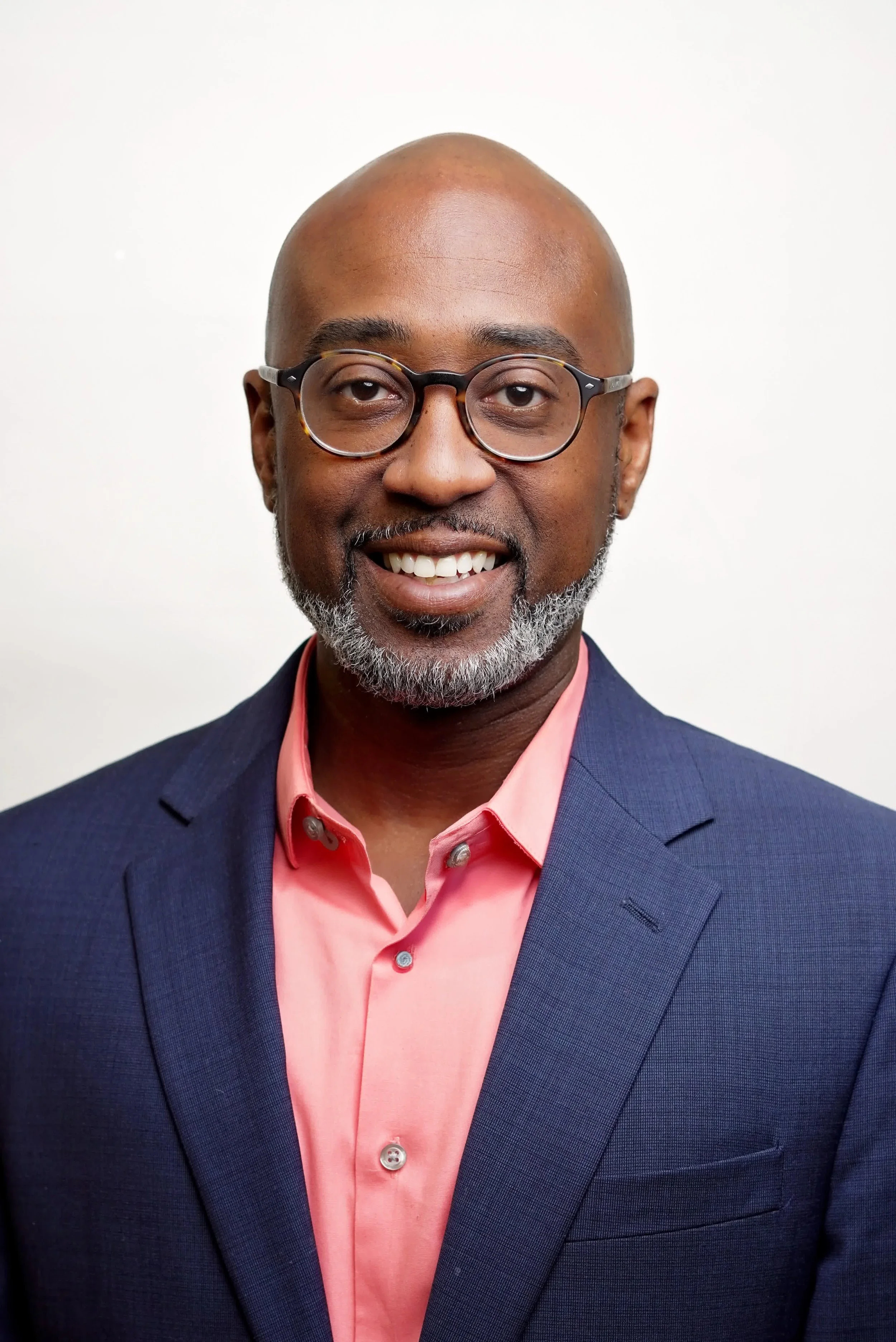 A professional headshot of a middle-aged African American man with a bald head, glasses, a gray beard, smiling, wearing a navy blazer and a pink shirt, standing against a plain white background.