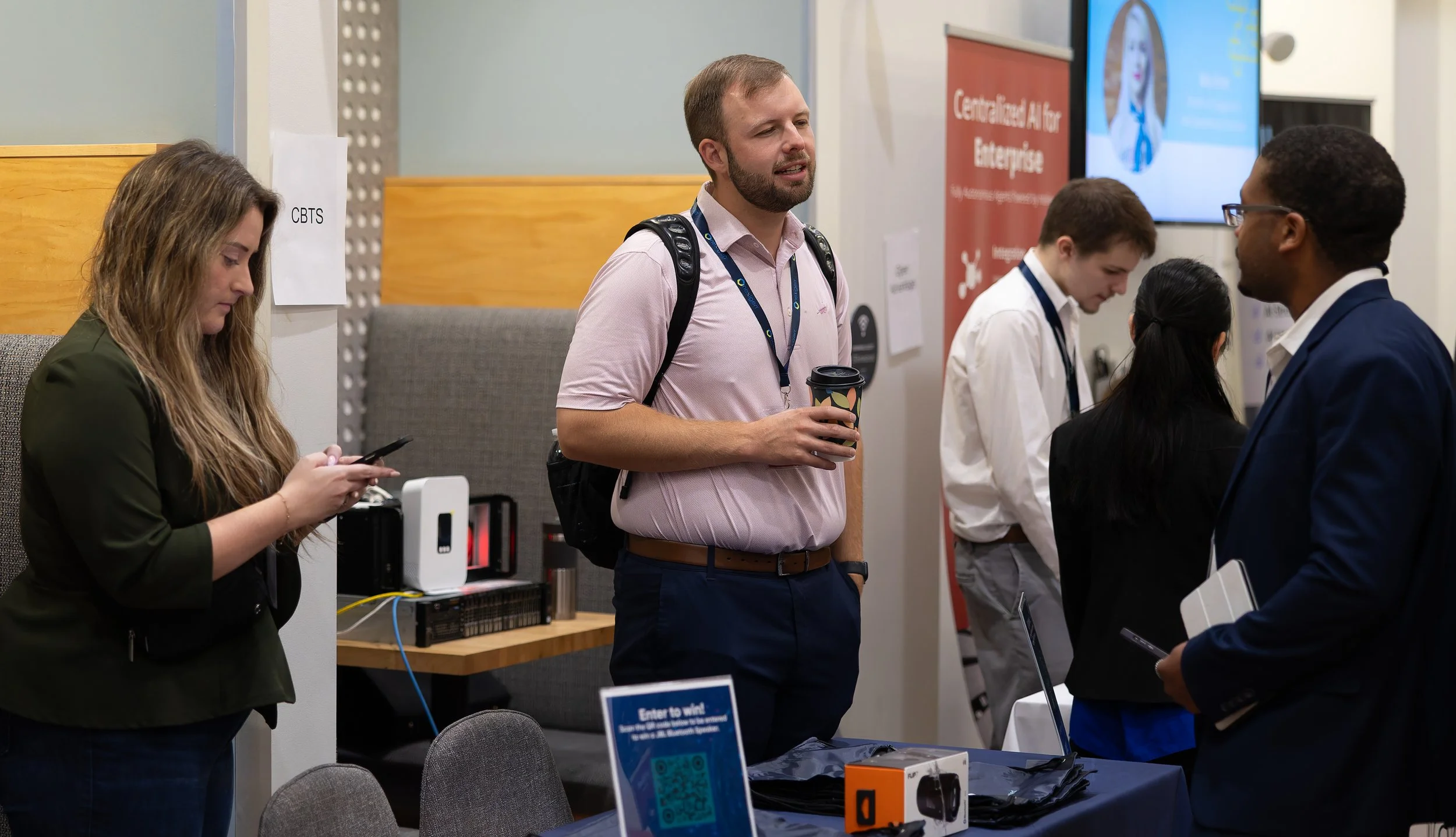 Group of five people at a professional conference, engaging in conversation and using electronic devices, with banners and a presentation screen in the background.