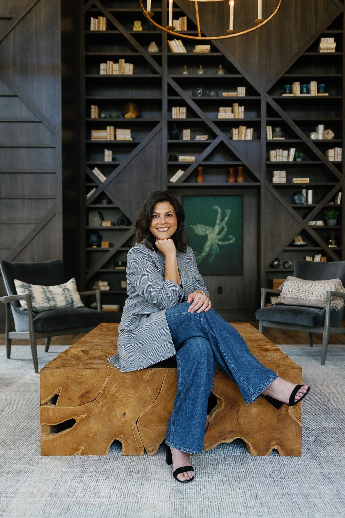 A woman sitting on a large wooden coffee table in a modern, decorated room with a dark wooden bookshelf behind her, two armchairs with pillows, and a framed abstract painting, smiling at the camera.