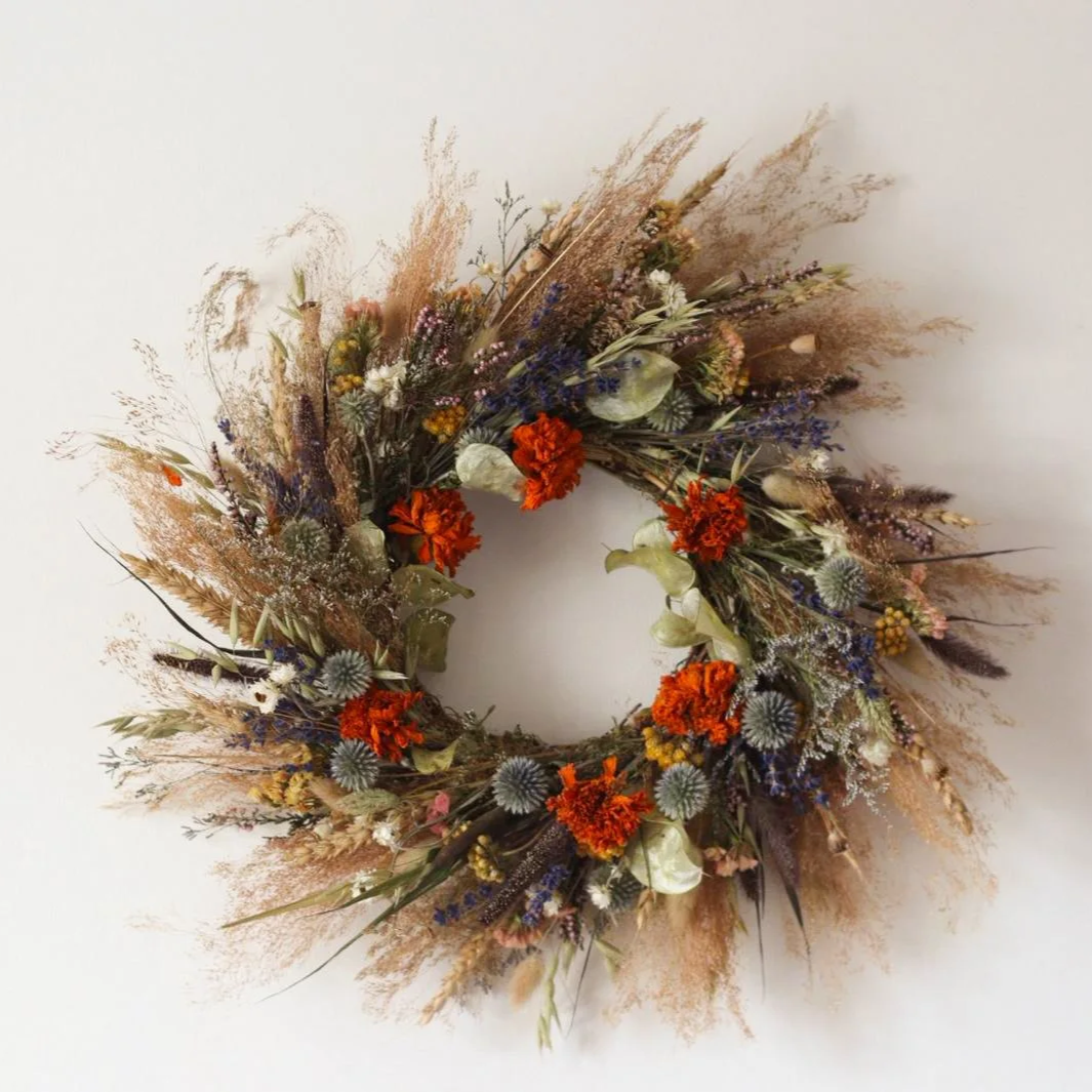 A dried floral wreath with orange, purple, white, and beige flowers and grasses, arranged in a circular shape on a white background.