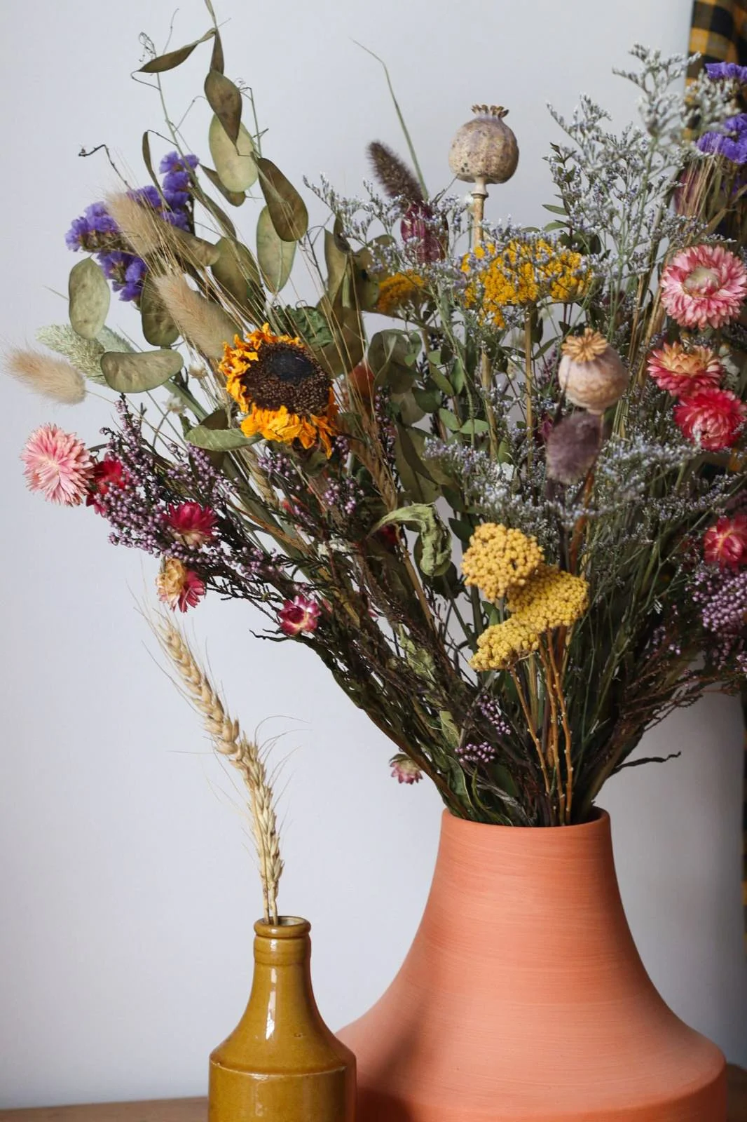 Dried flower arrangements in a large terracotta vase and a small mustard-colored bottle on a wooden surface.