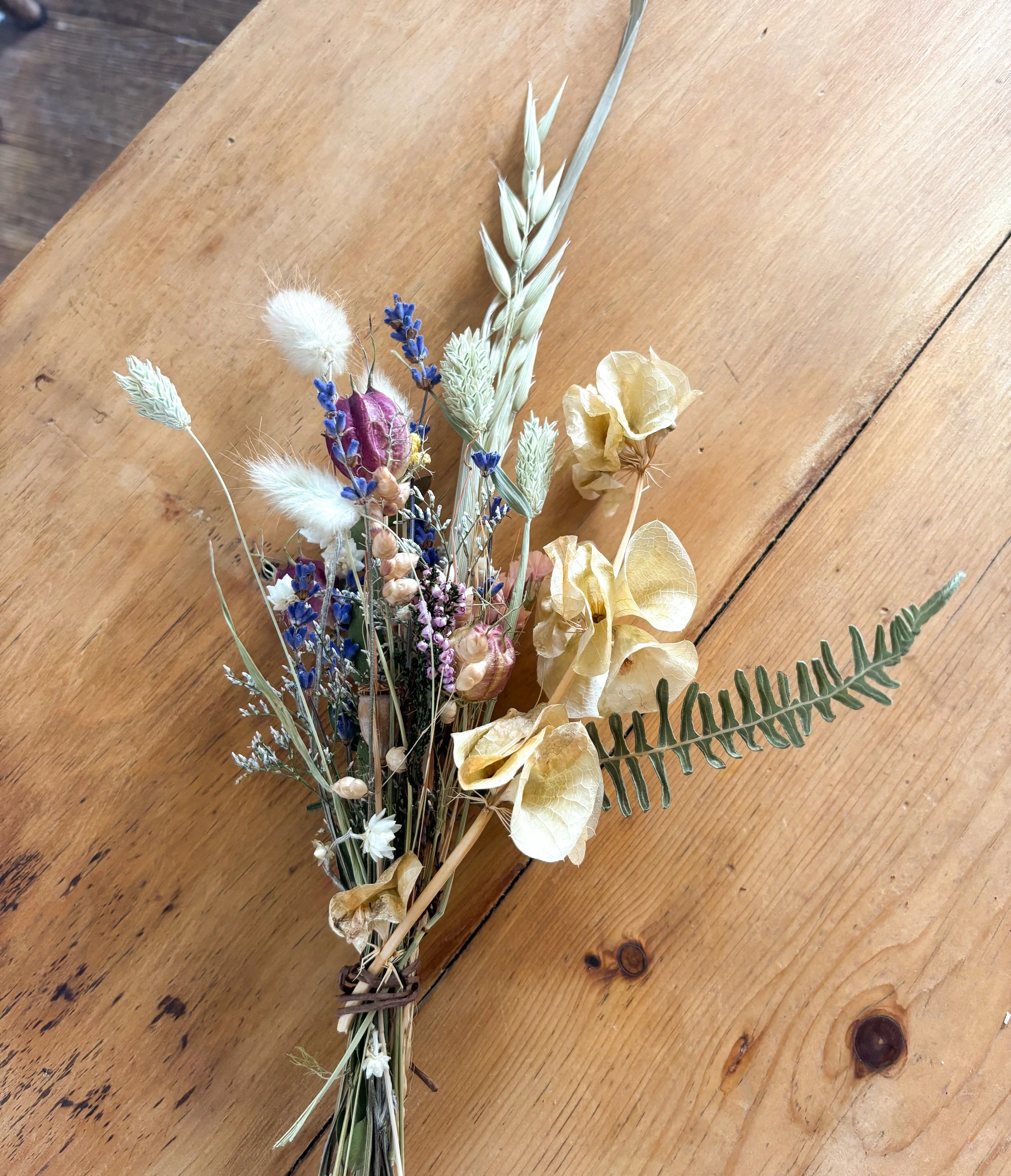 A bouquet of dried flowers including ferns, lavender, and various seed pods, placed on a wooden surface.