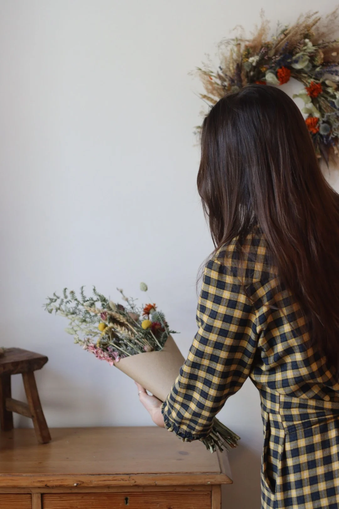 A woman with long dark hair wearing a yellow and black checkered shirt holding a bouquet of dried flowers inside a room with a wooden table and a floral wreath on the wall.