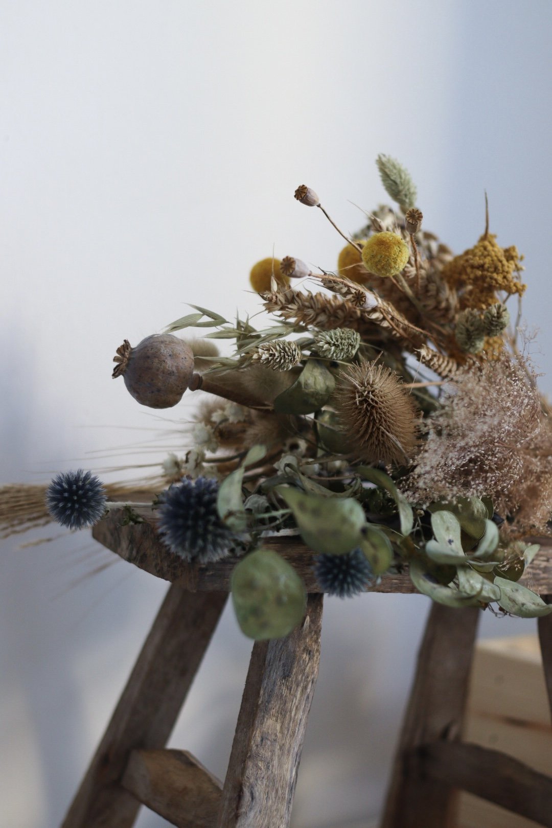 A dried bouquet of various flowers and seed pods resting on a rustic wooden stool against a plain background.
