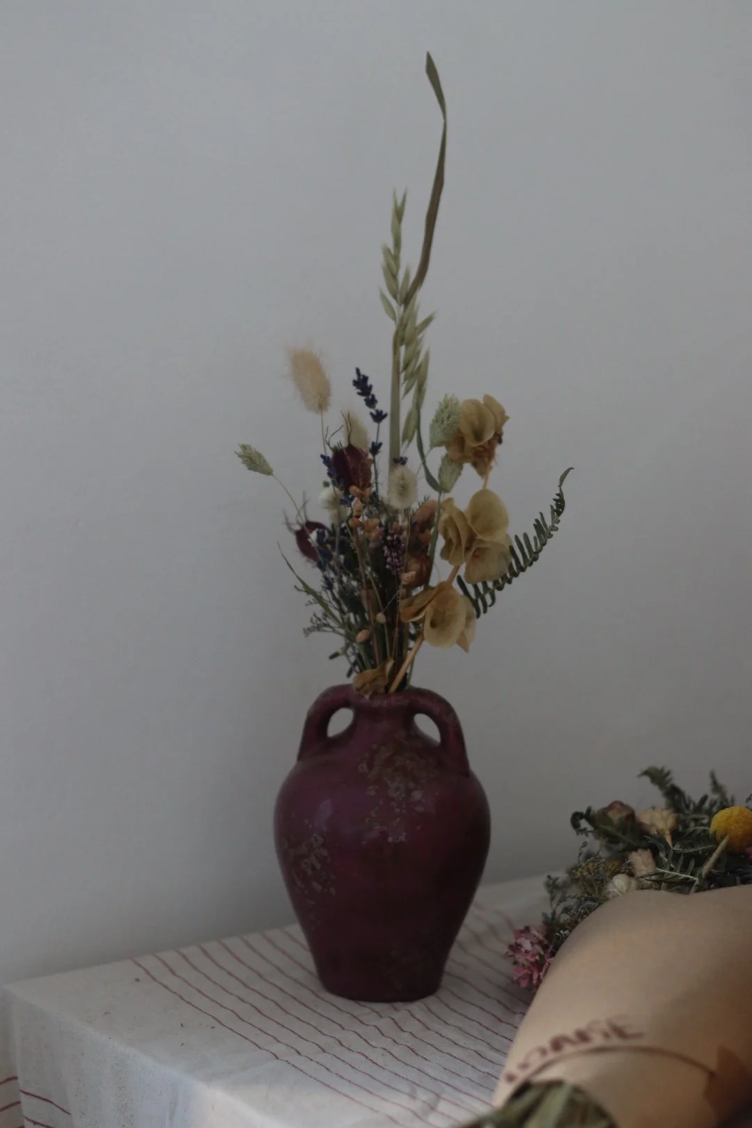 A maroon ceramic vase with dried wildflowers, including grasses, lavender, and beige roses, placed on a white table with a striped cloth against a plain light-colored wall.