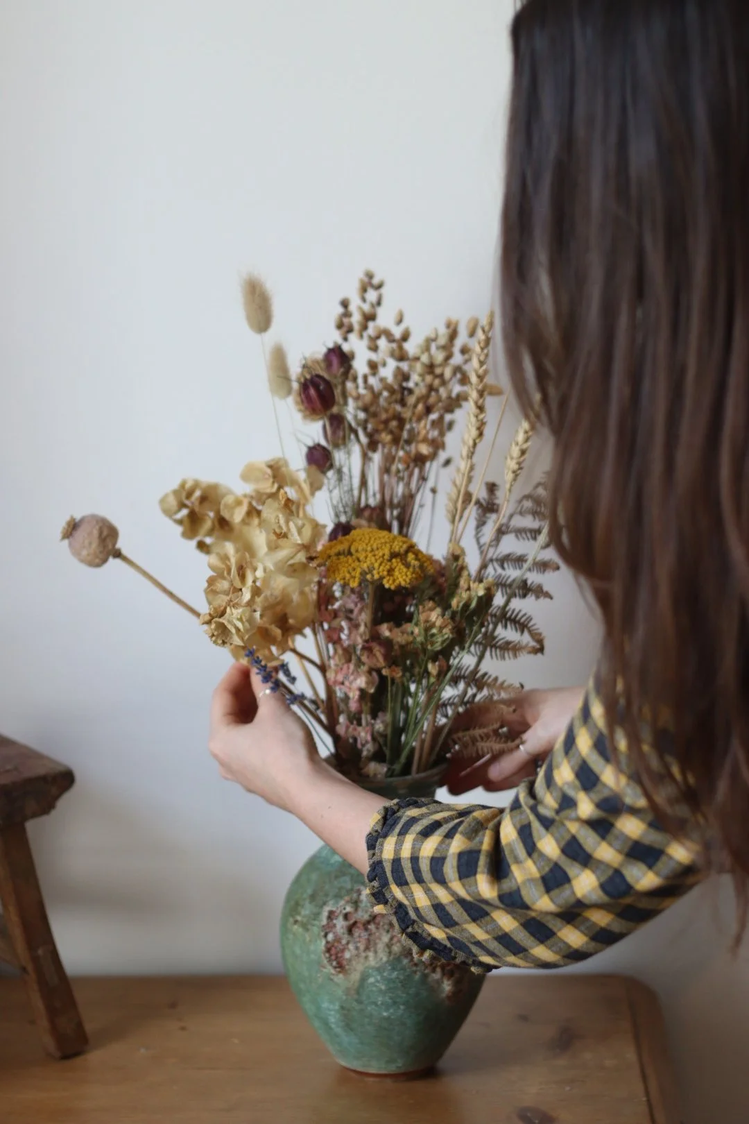 A person with brown hair wearing a yellow and black checkered shirt arranging a bouquet of dried flowers in a green ceramic vase on a wooden surface.