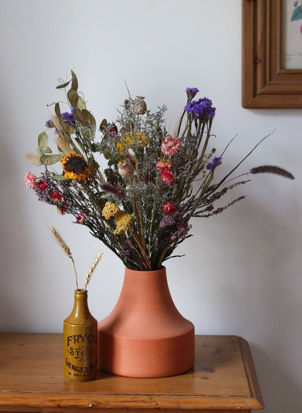 A bouquet of mixed dried flowers in a large, terracotta-colored vase on a wooden table, with a small vintage yellow bottle containing a single dried wheat stalk beside it.