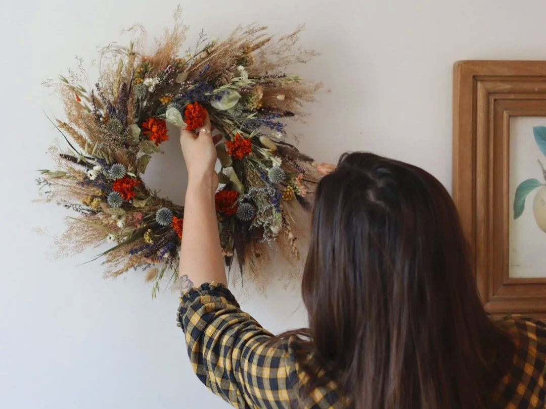 A woman is hanging a dried flower wreath on a wall.