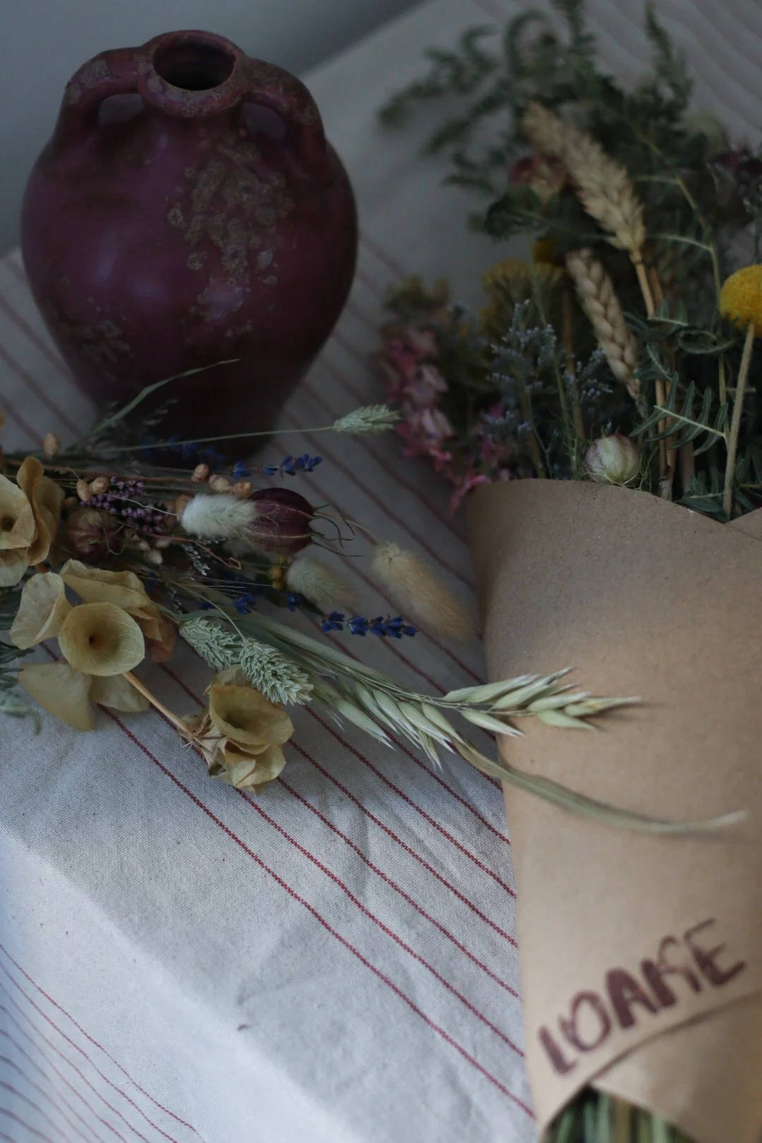 A rustic still life featuring an antique purple-brown ceramic jug and a bouquet of dried flowers on a white cloth with red stripes.