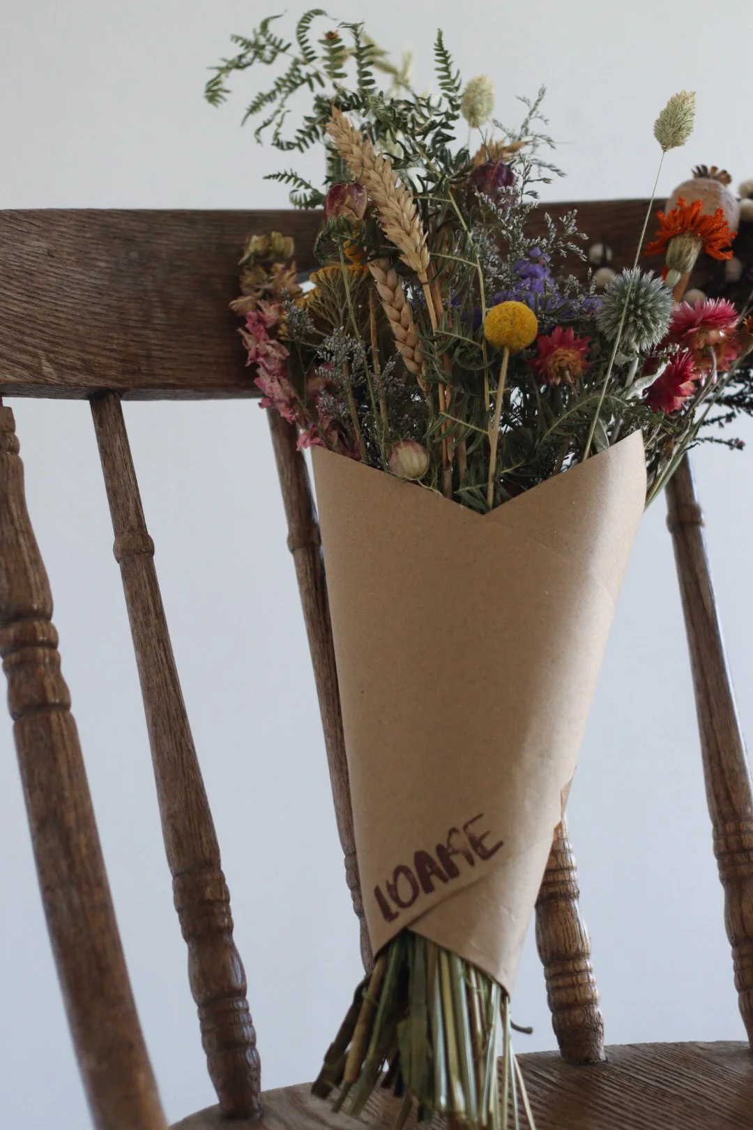 A bouquet of dried wildflowers wrapped in brown paper with the word 'LOAFE' written on it, resting against a wooden chair.