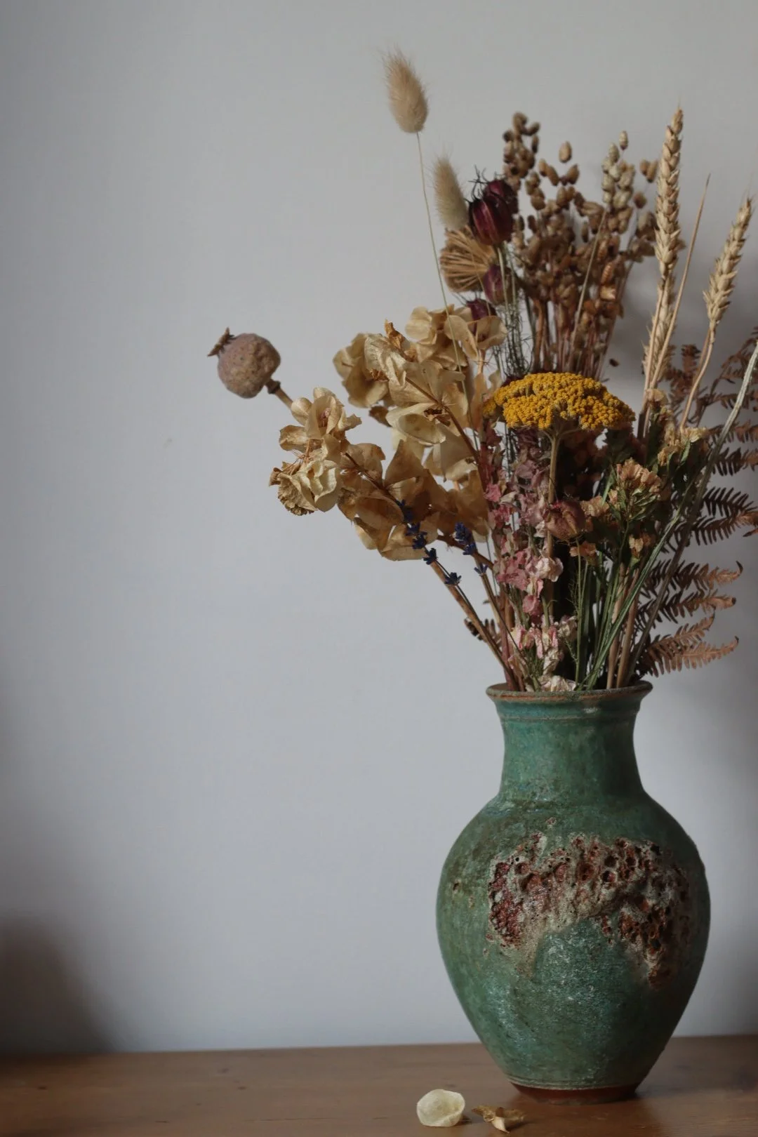 A rustic ceramic vase with a weathered green finish holding a bouquet of dried flowers, including ferns, hydrangeas, and various grasses, placed on a wooden surface against a plain white wall.