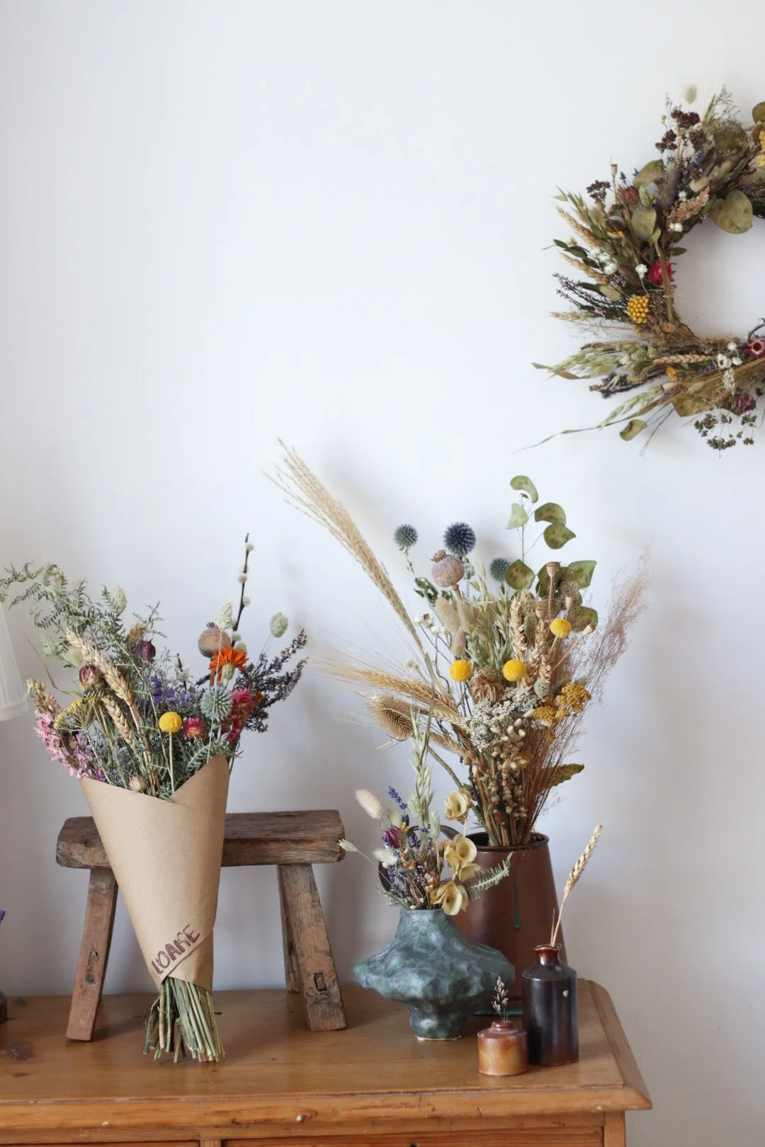 Three vases filled with dried flowers and plants on a wooden table, with a wreath of dried flowers hanging on the wall behind.