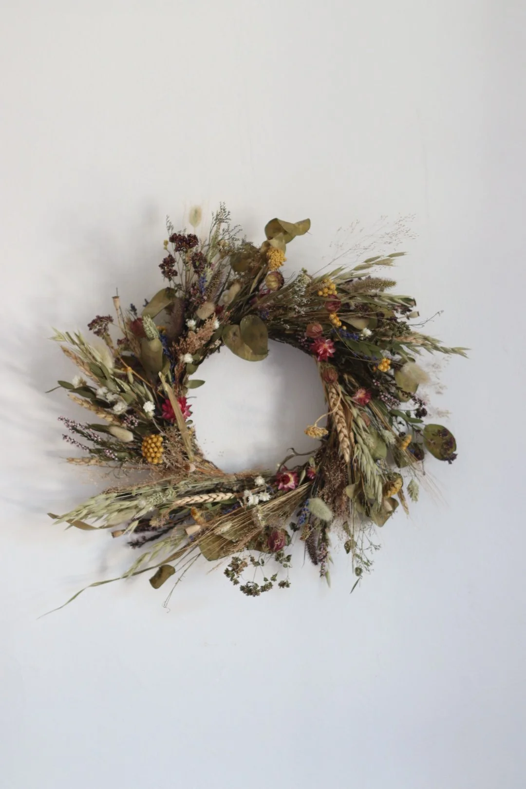 Dry floral wreath with various dried flowers and grasses hanging on a white wall.