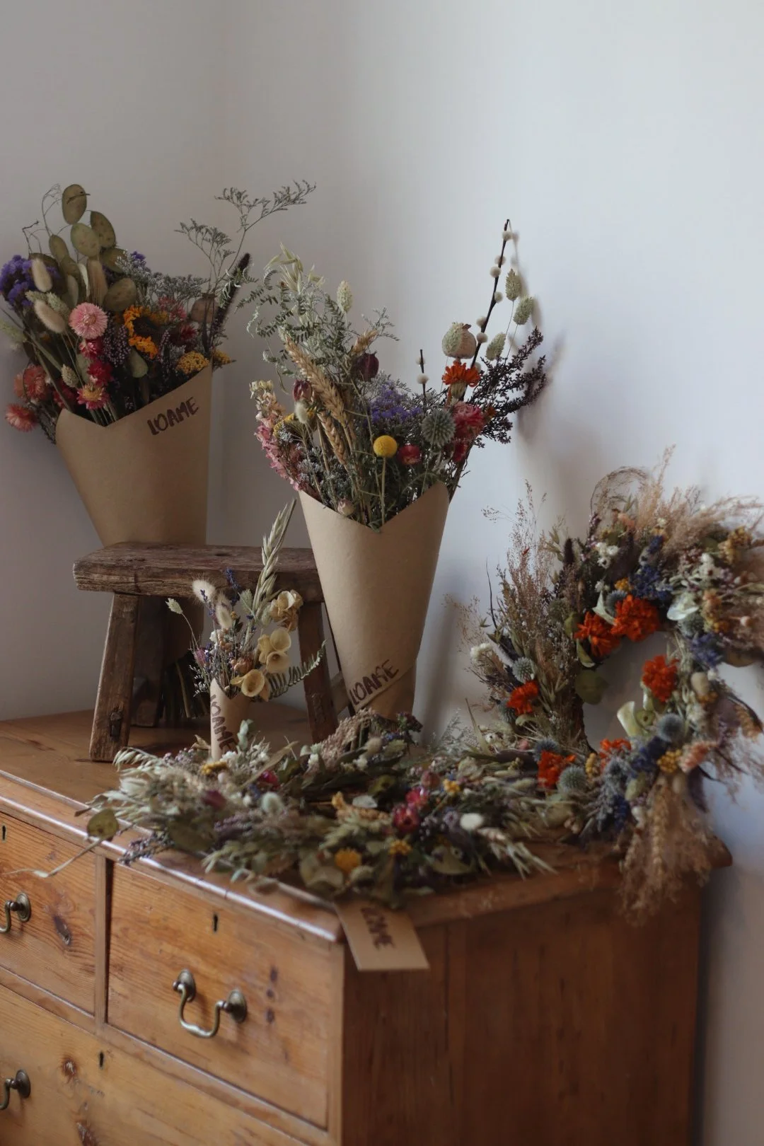 Several bouquets of dried flowers in paper wrapping, placed on and around a wooden table, with a small rustic stool and a flower wreath on the table.