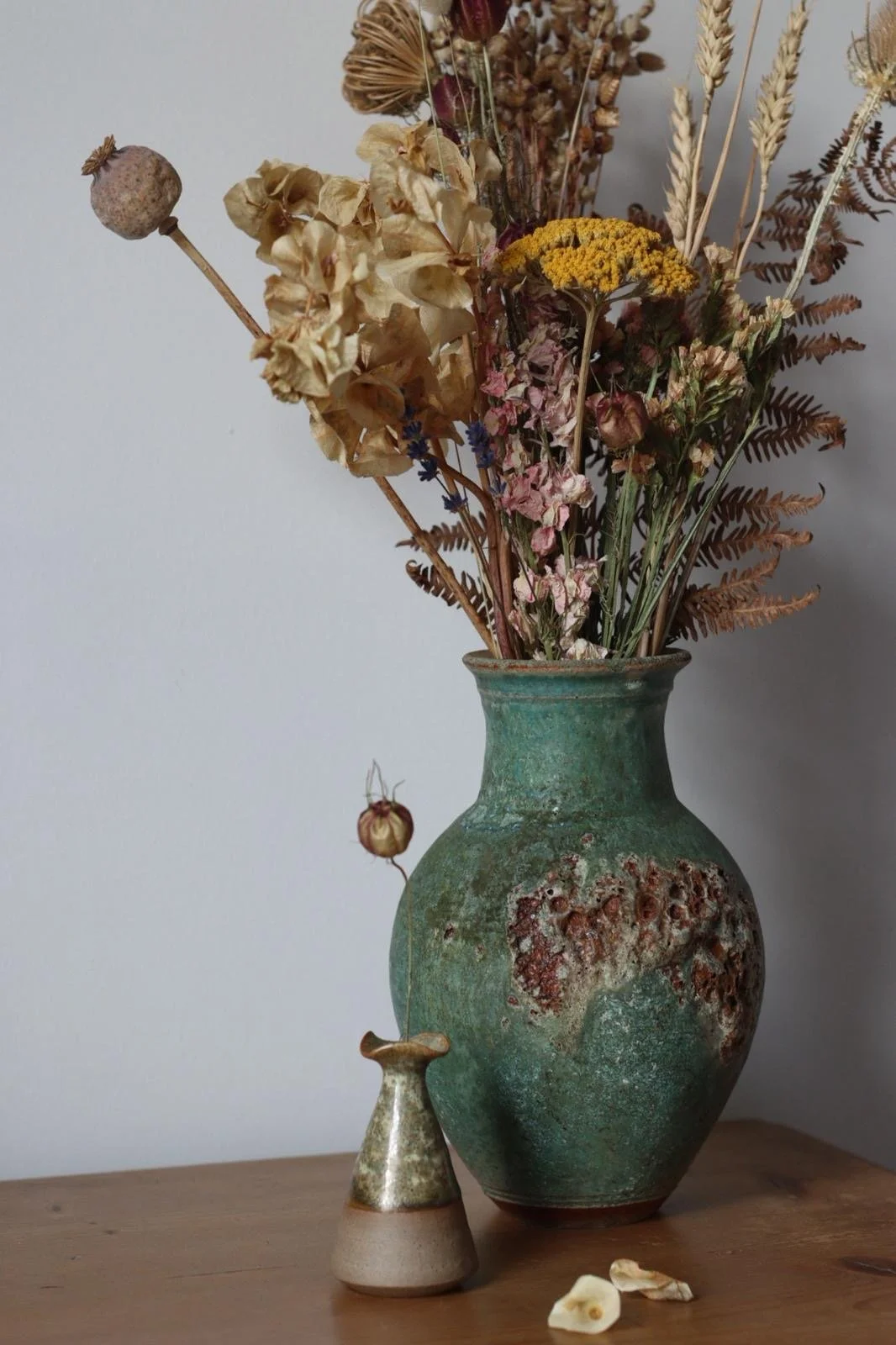 A ceramic vase with a textured, weathered green surface holding a bouquet of dried flowers and foliage, placed on a wooden surface against a plain wall.