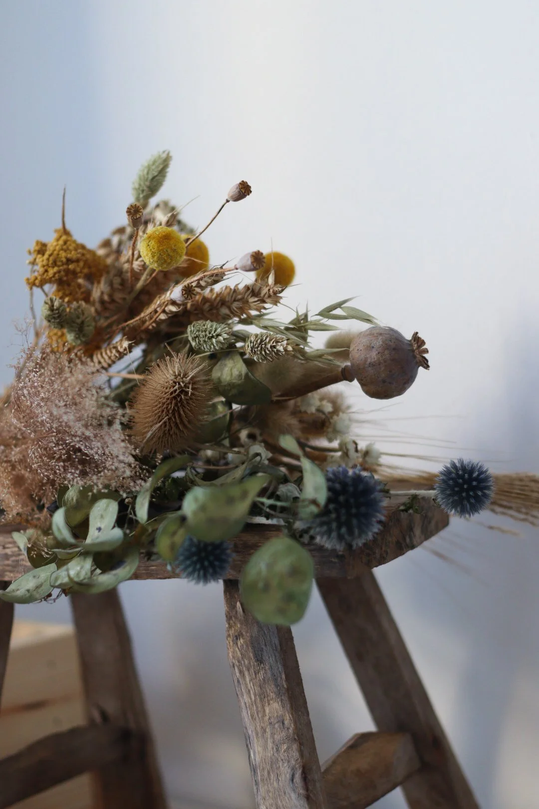 A rustic dried flower arrangement on a wooden stool, featuring various dried flowers, seed pods, and grasses in natural tones.
