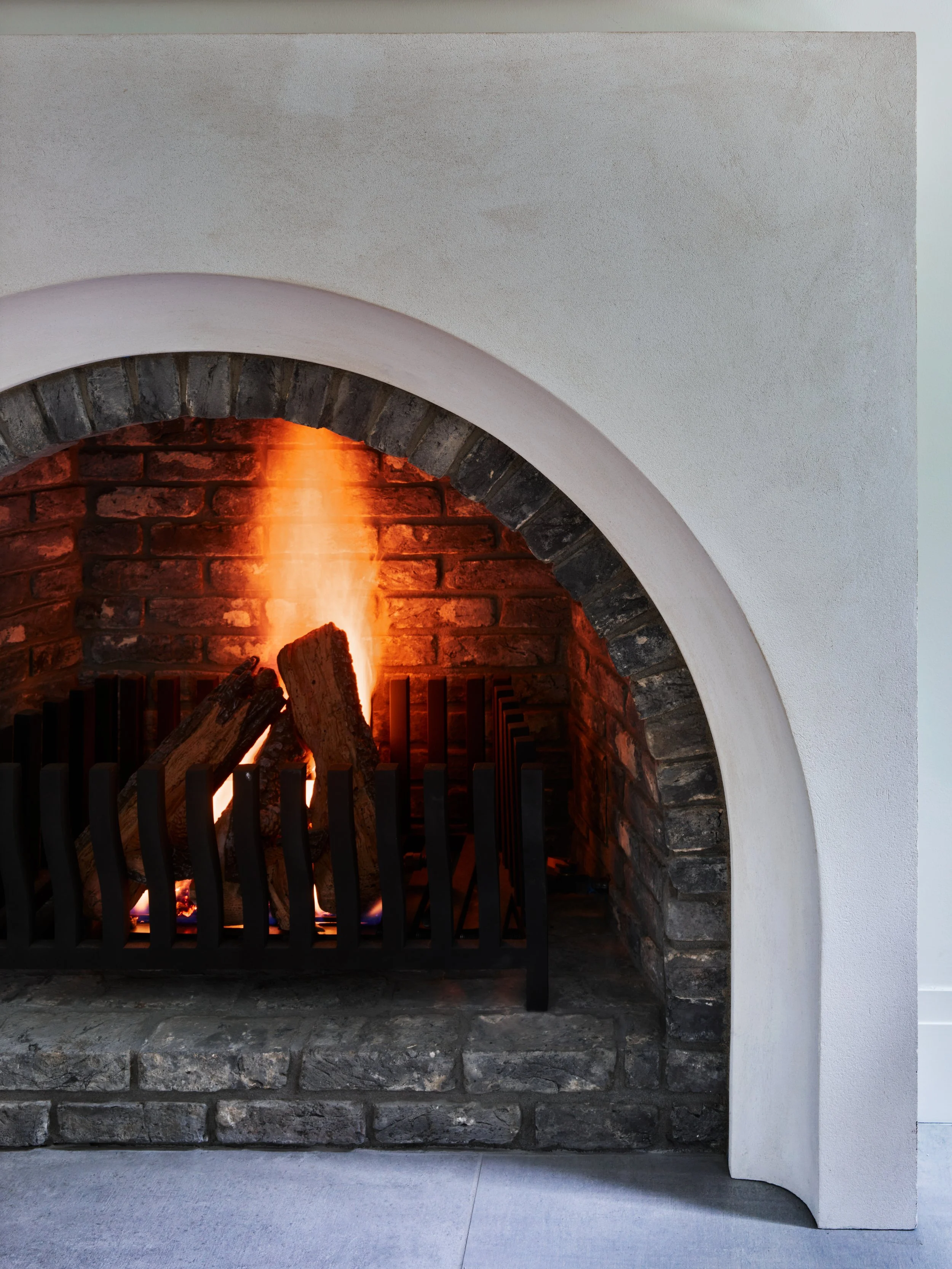 Modern indoor fireplace with a brick interior, a fire burning with orange flames, and a black metal grate. The fireplace is framed by an arched brick opening and a smooth, white wall.
