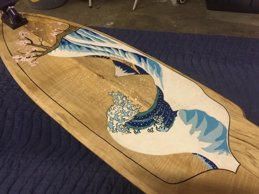 A wooden surfboard with a painted design of a large wave, a flowering tree branch, and a mountain in the background, resting on a blue quilted surface.