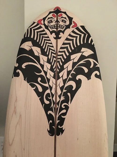 A surfboard with a traditional Polynesian black and red tattoo design, featuring a stylized face at the top and intricate patterns down the board's length.