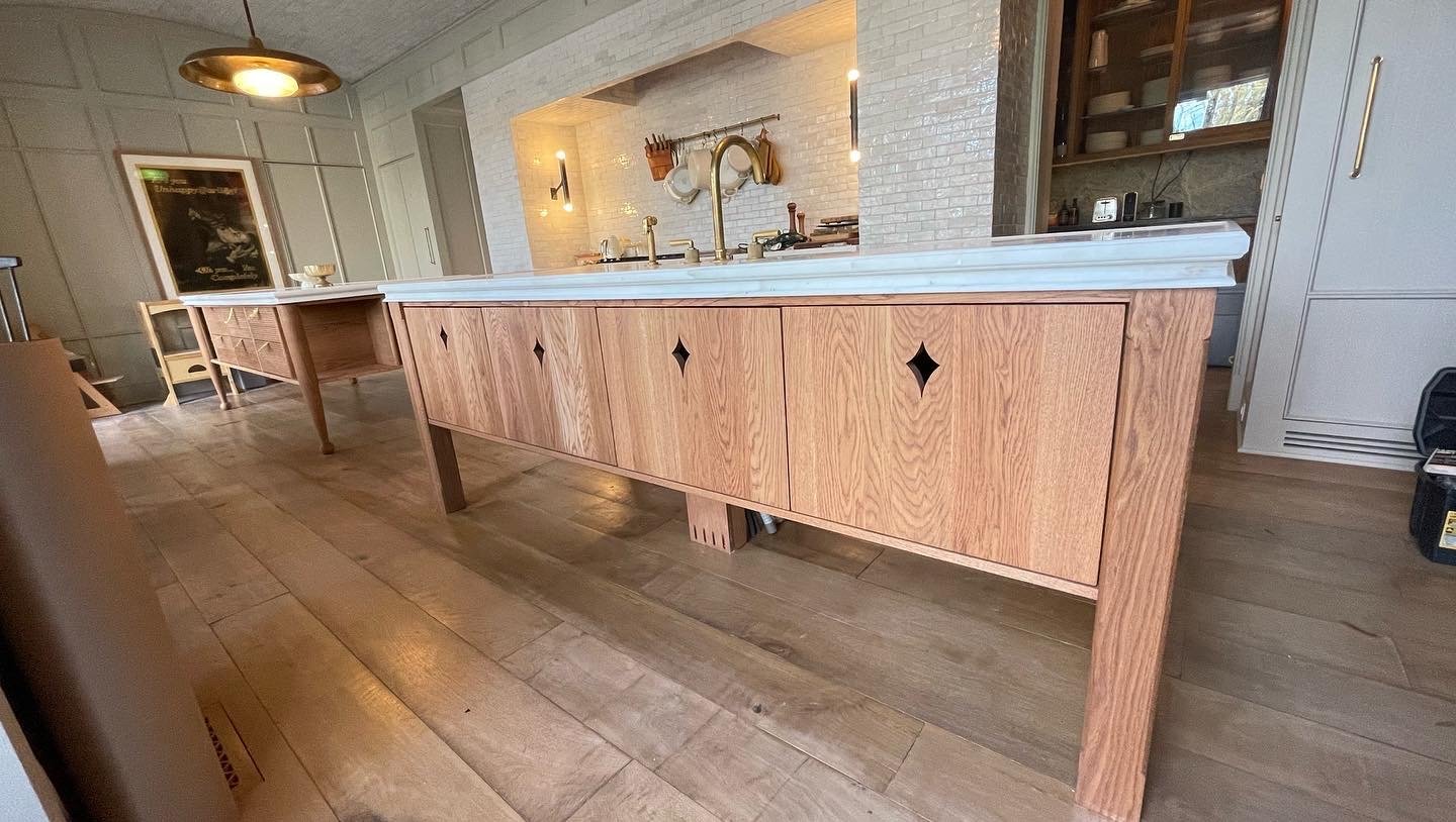 Kitchen with wooden island in the center, white marble countertop, brass fixtures, and wood cabinetry, with a brick wall and open shelves in the background.