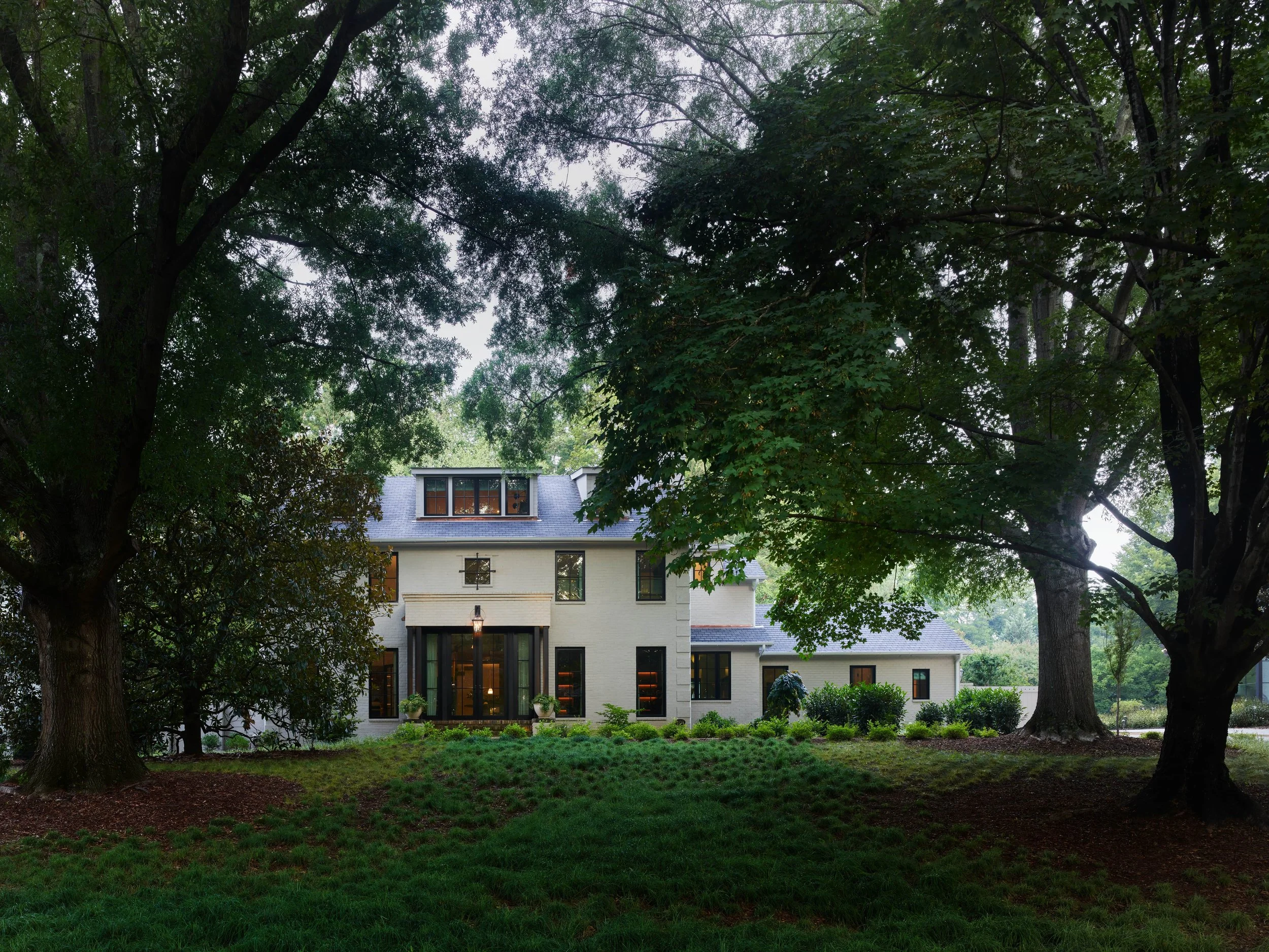 A large house with big windows is surrounded by tall trees and green grass in a lush yard.