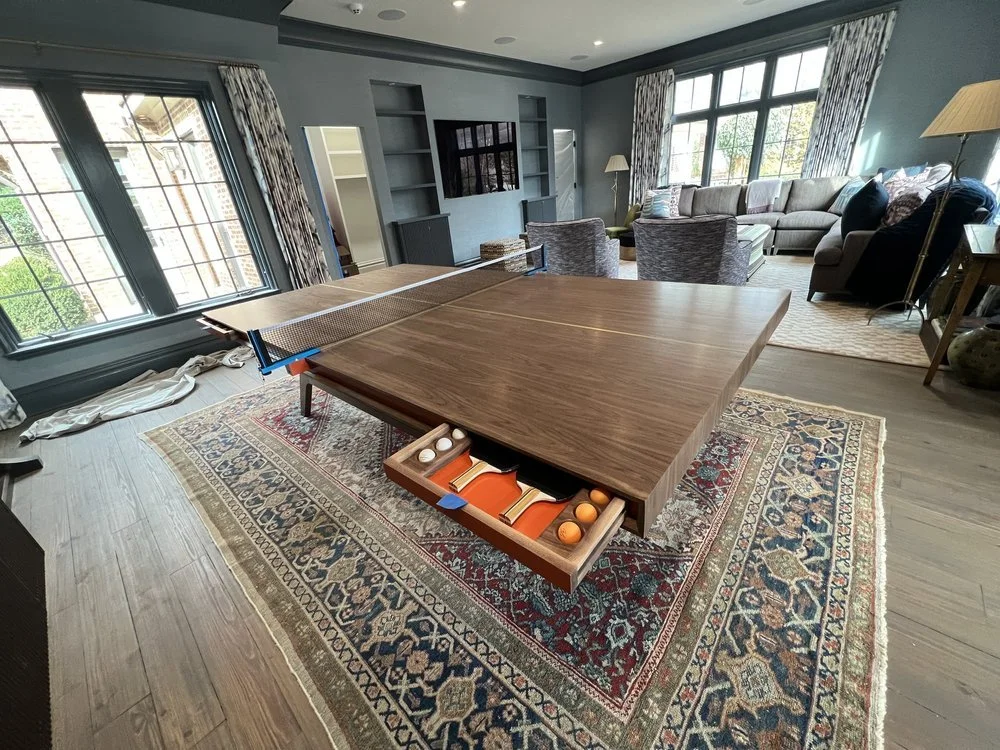 Living room with large windows, a ping pong table with a wooden top, and a patterned area rug. There is a sectional sofa with pillows, a side table with a lamp, and a TV mounted on the wall.