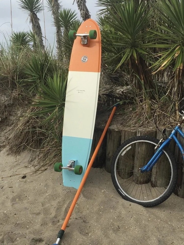 A colorful skateboard is leaning against a wooden post on a sandy beach, with a blue bicycle partly visible next to it and tropical plants in the background.