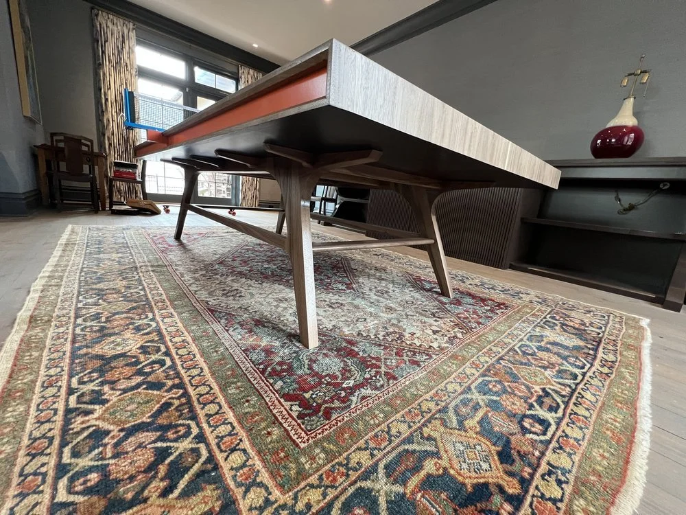 Zoomed-out view of a wooden dining table set on a detailed, ornate area rug in a living room, with windows and curtains in the background.