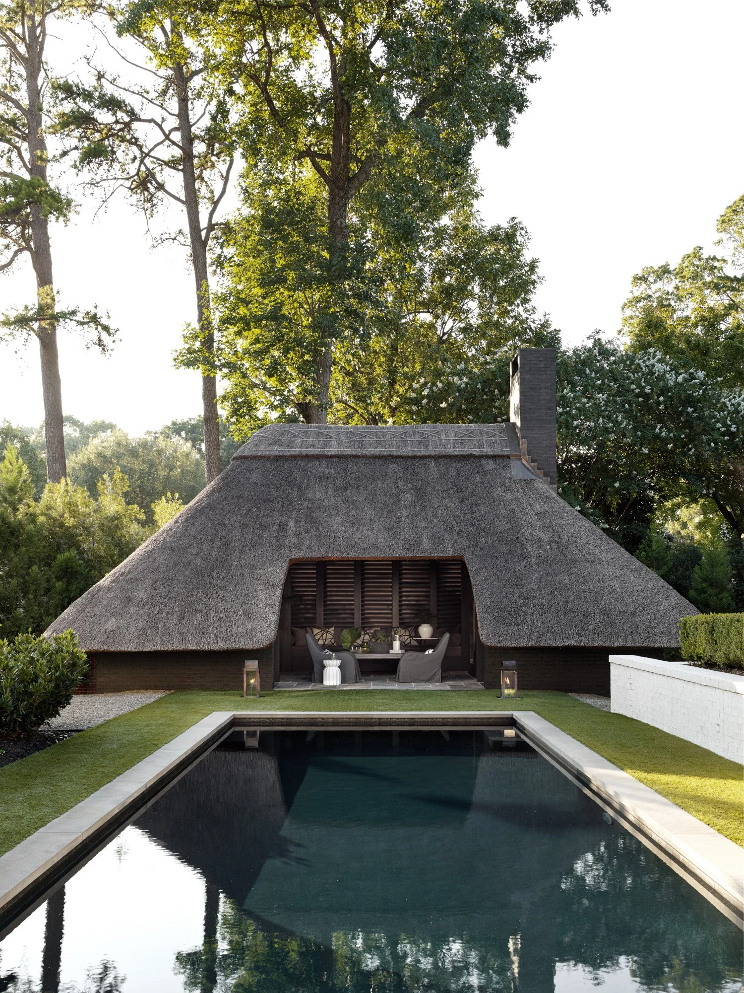A backyard with a rectangular reflecting pool, a thatched-roof cabana with outdoor seating, surrounded by green grass, shrubs, and tall trees, during daylight.