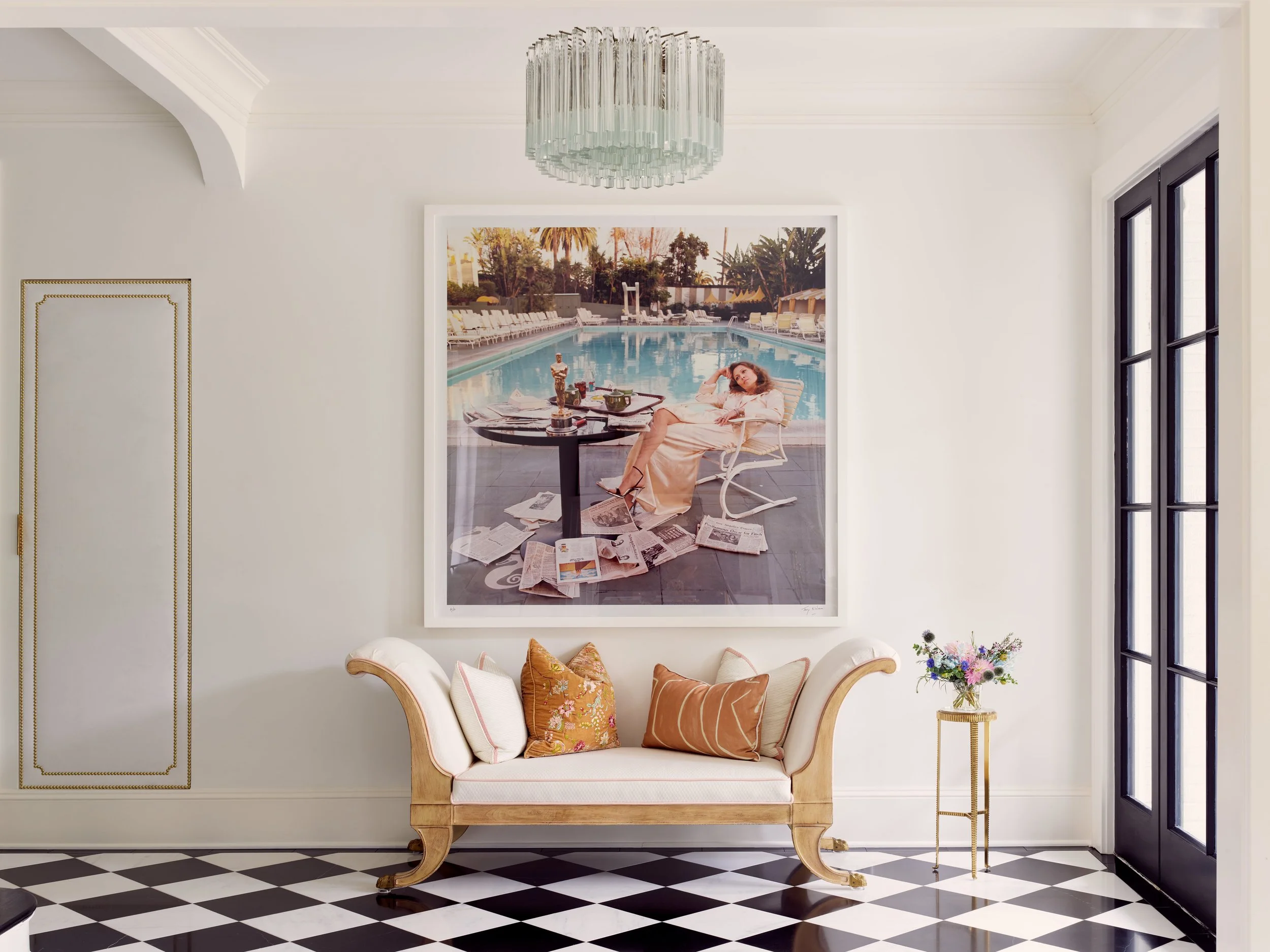 Interior of a living room with black and white checkered floor, a white sofa with rust, pink, and floral pillows, a small round side table with a vase of flowers, and a large framed photograph of a woman relaxing by a swimming pool on the wall behind