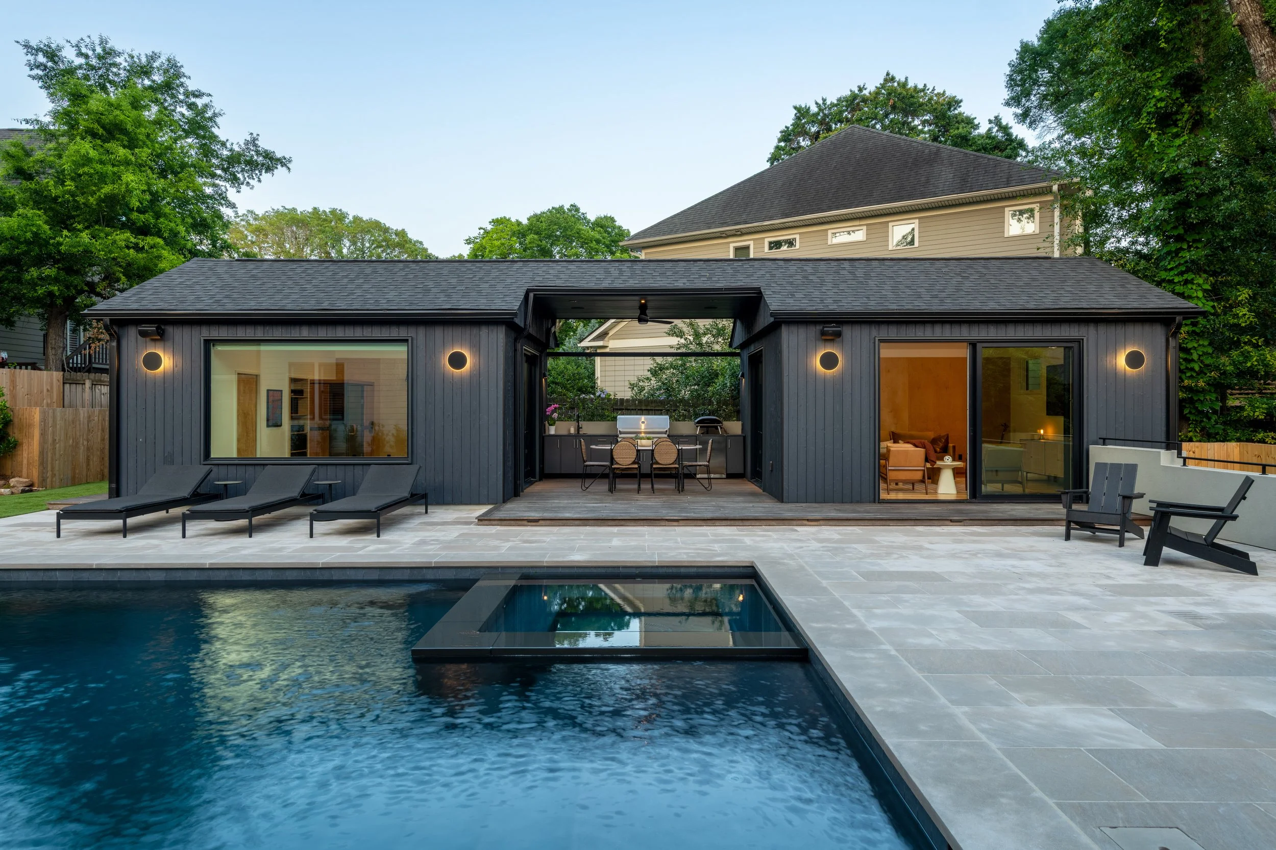 A modern backyard with a swimming pool, a black wooden pool house with large windows, outdoor lounge chairs, and a patio table with chairs. There are green trees and a neighboring house in the background.