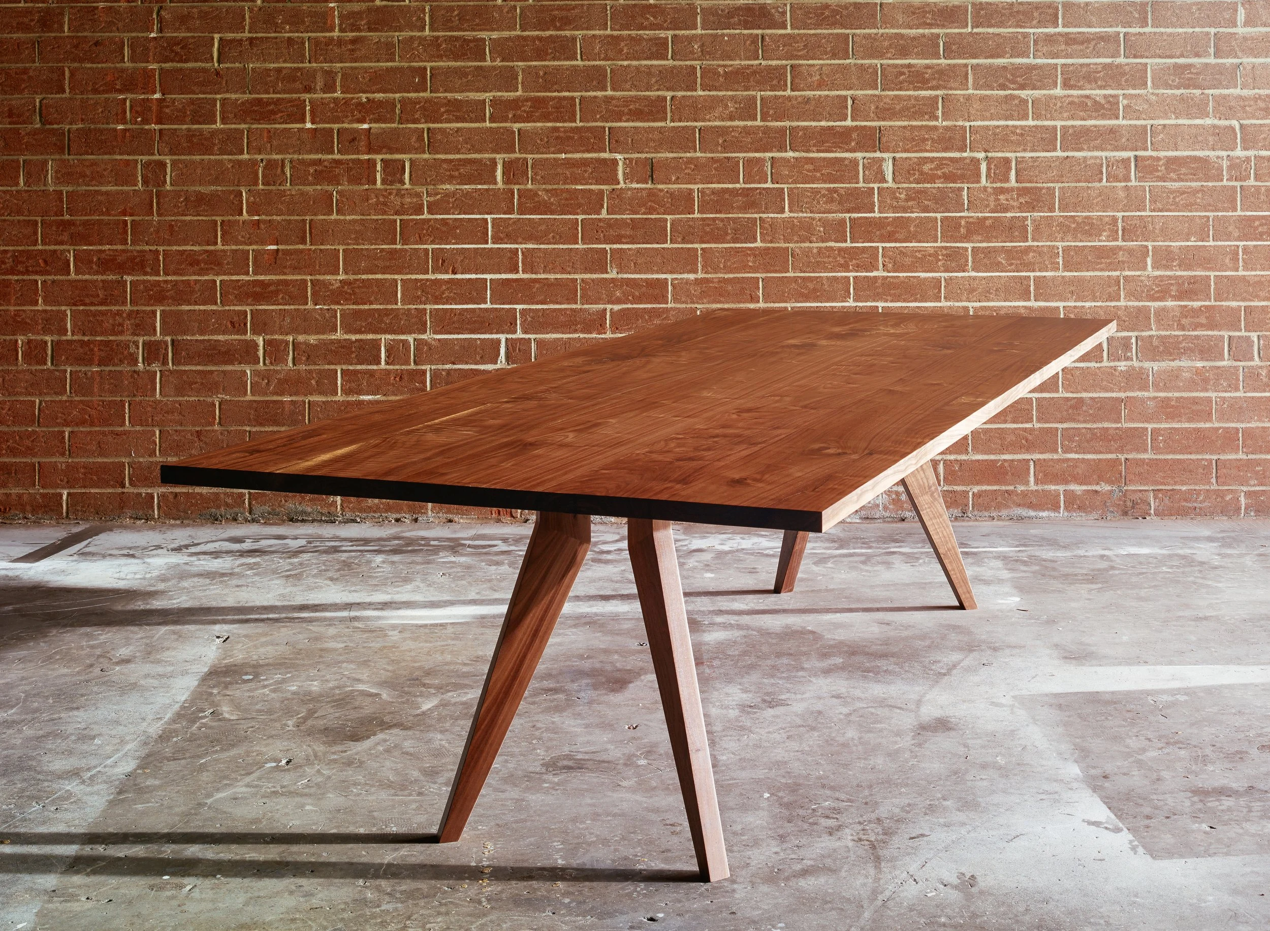 A wooden table with angled legs is placed against a red brick wall in an unfinished or industrial space with concrete floors.