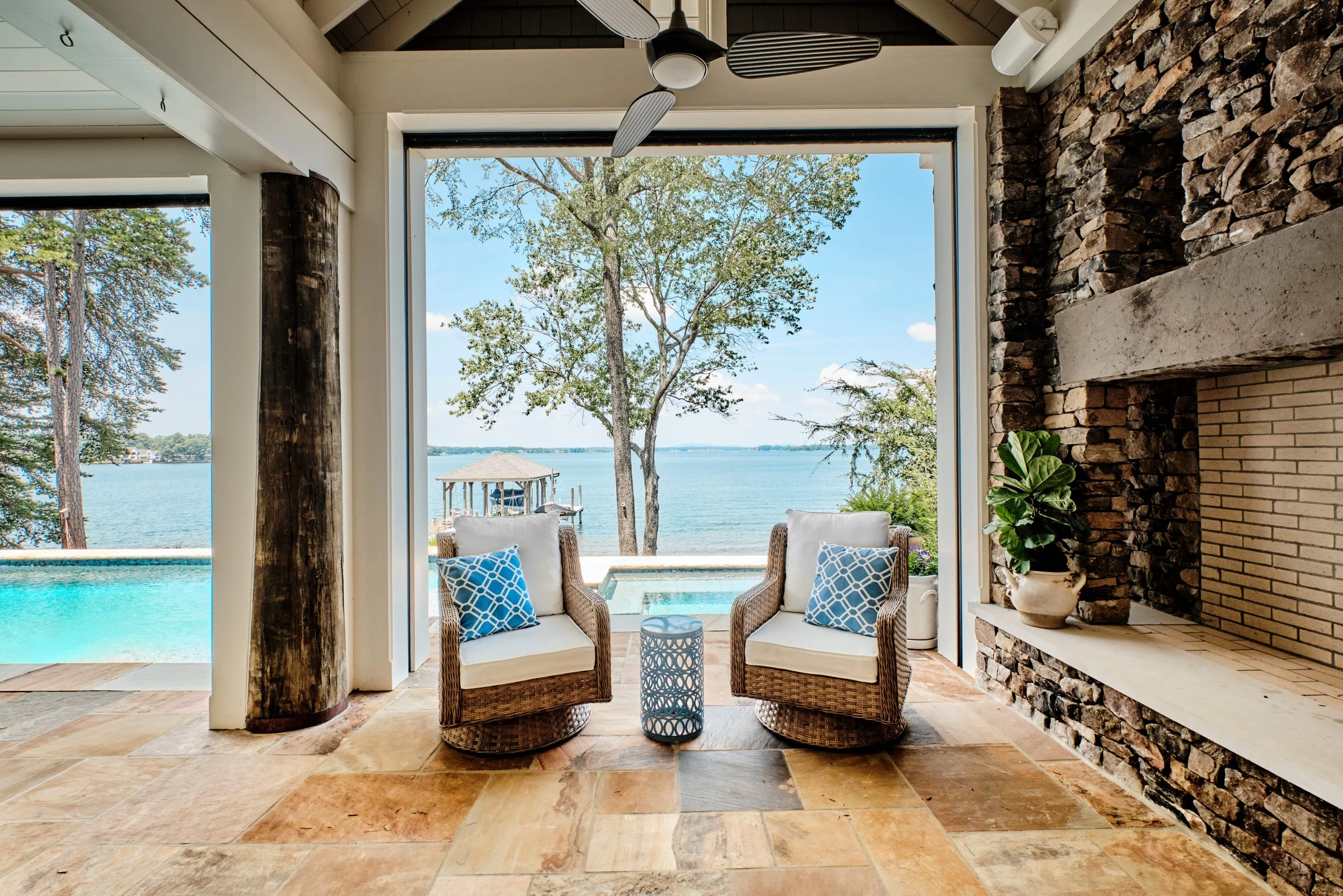Living room with a large window view of a lake, trees, and a gazebo. Two wicker chairs with white cushions and blue patterned pillows are in front of the window, accompanied by a small round side table. The room has stone and brick accents, a ceiling