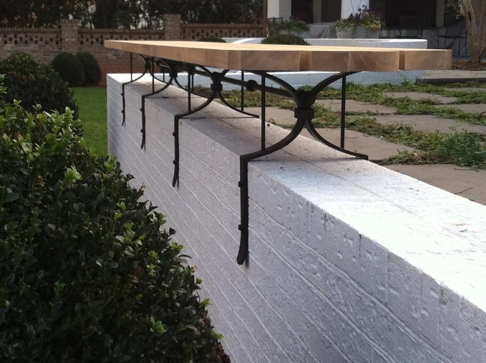 Close-up of a white brick wall with decorative black steel brackets that support a wooden planter box on top, set in a garden with bushes and paved pathway.