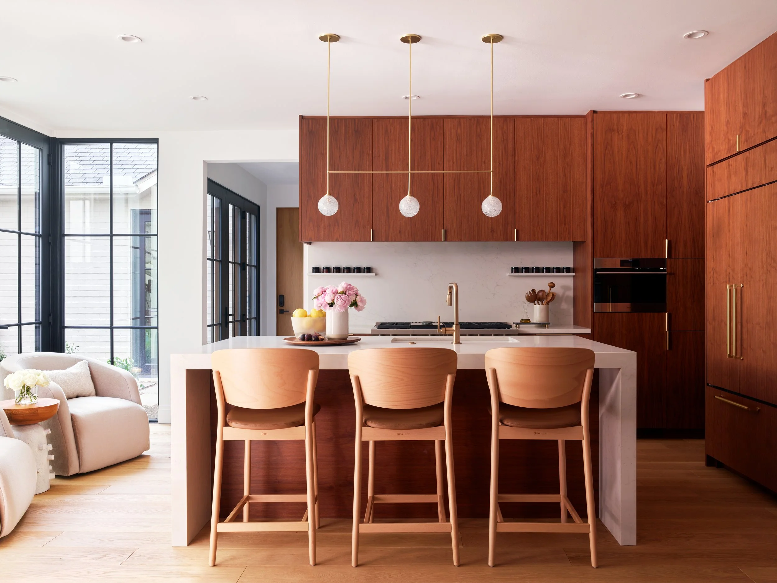 Modern kitchen with wood cabinetry, hanging pendant lights, white island with a gold faucet, and large windows to the left.