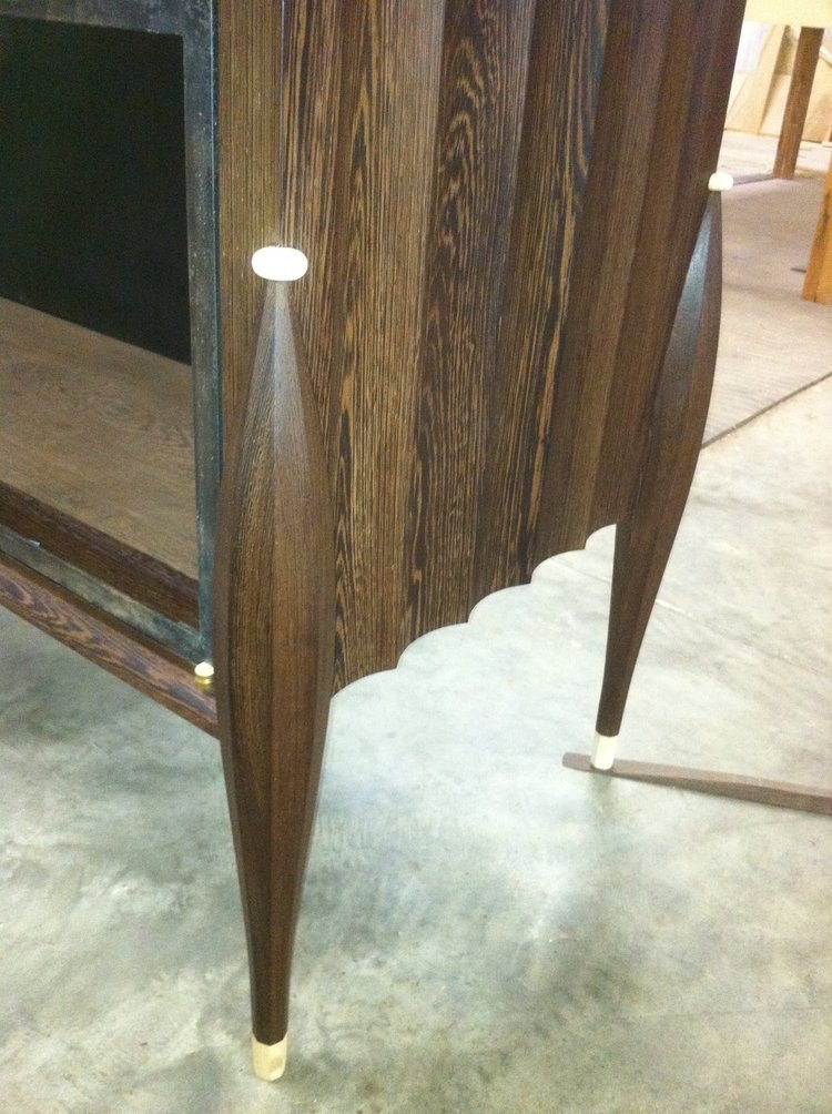 A close-up of a wooden cabinet with a dark brown finish and tapered legs with gold-colored caps, standing on a concrete floor.