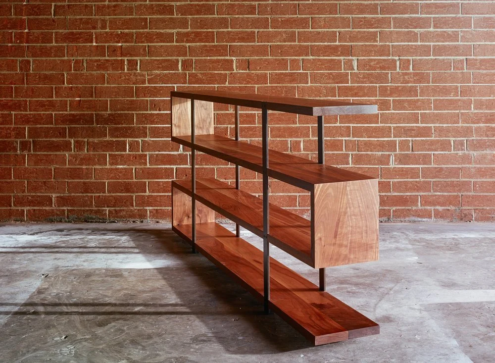 A modern wooden bookshelf with metal supports against a brick wall and concrete floor.