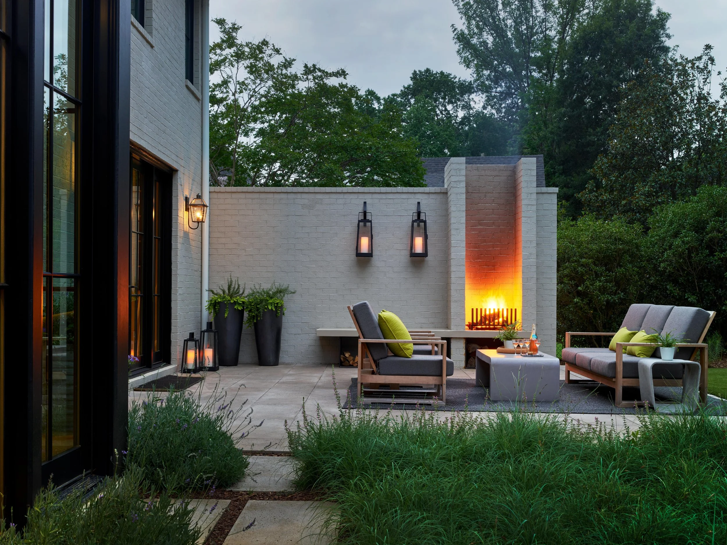 A cozy outdoor patio with gray seating, wooden furniture, and a fireplace, surrounded by lush greenery and evening lighting.