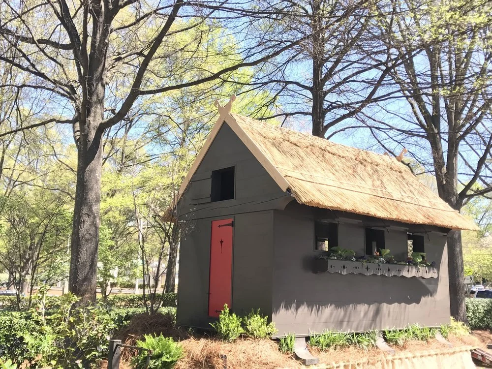 A miniature house with a gray exterior, red door, small windows, and a thatched roof surrounded by trees and greenery.