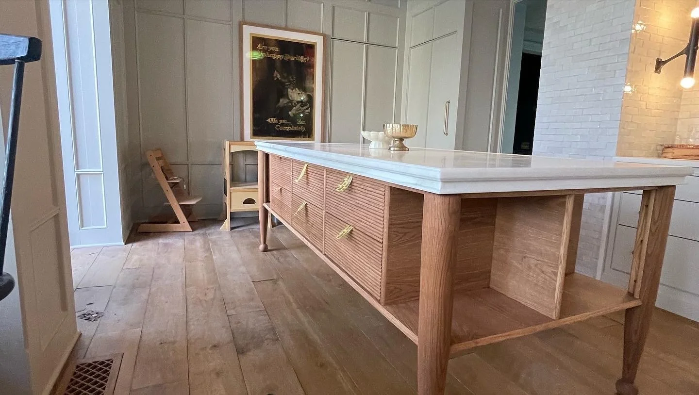 A modern kitchen island with a white marble top and a wooden base with drawers and open storage space, situated in a room with wooden flooring and white-paneled walls.