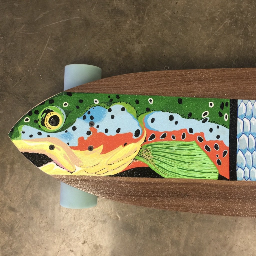 Colorful skateboard deck with a painted rainbow trout fish, featuring green, blue, yellow, orange, and black spots, mounted on a wooden board with a light blue wheel visible at the top left.