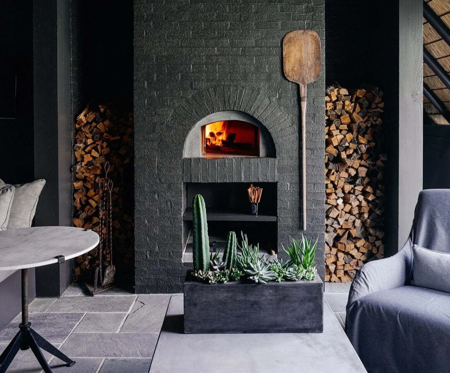 Indoor scene featuring a modern black brick fireplace with a fire burning inside. Two stacks of firewood are stored on either side of the chimney. A wooden pizza peel leans against the wall next to the fireplace. In front of the fireplace, a rectangu