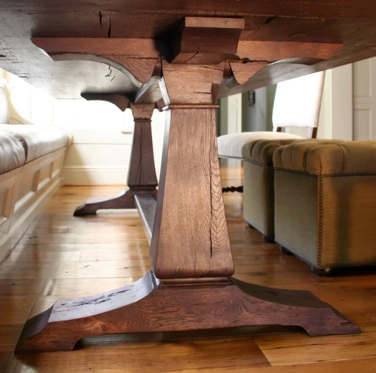 Close-up view of a sturdy wooden dining table leg and base, with part of a cushioned bench and armchair in a well-lit room with wooden flooring.
