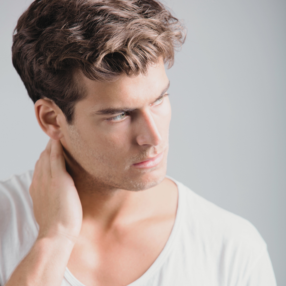 A young man with brown hair and blue eyes, wearing a white t-shirt, touching the back of his neck, with a contemplative expression on his face.