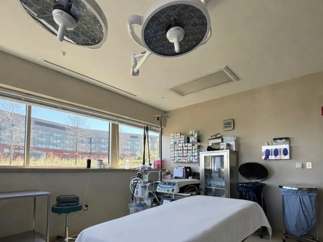 Empty hospital operating room with surgical lights, a bed covered with white sheets, medical equipment, and a large window with a view of a building and trees outside.