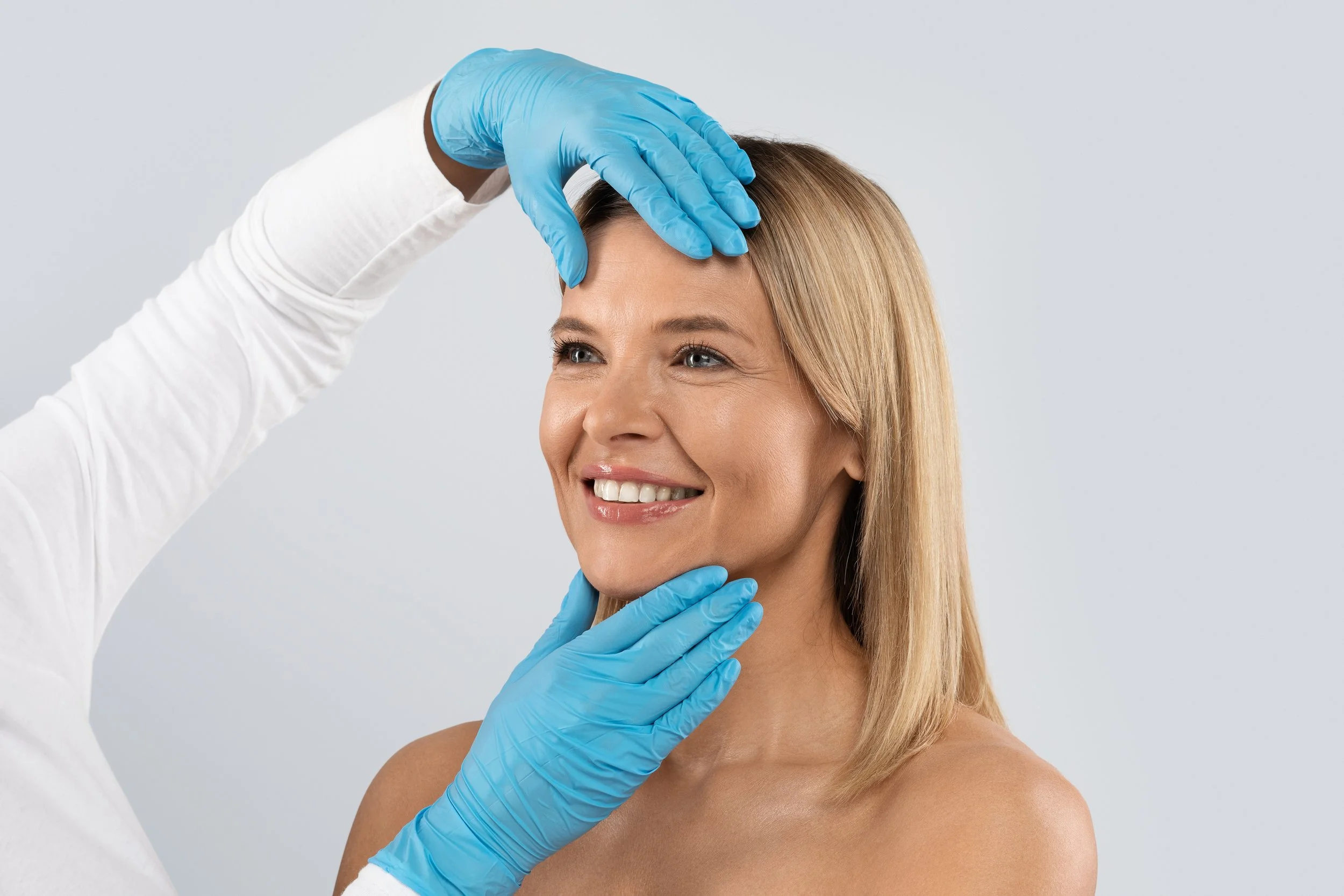 A woman receives a cosmetic or dermatological examination from a healthcare professional wearing blue gloves, with the healthcare worker checking her forehead and chin.