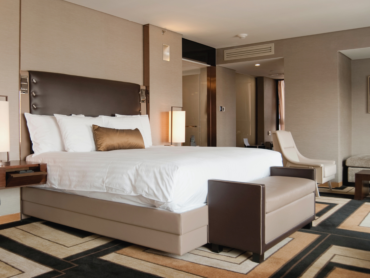 Luxurious hotel room with a large bed, white linens, and a gold throw pillow. The room has modern furniture, a patterned carpet, and soft lighting.