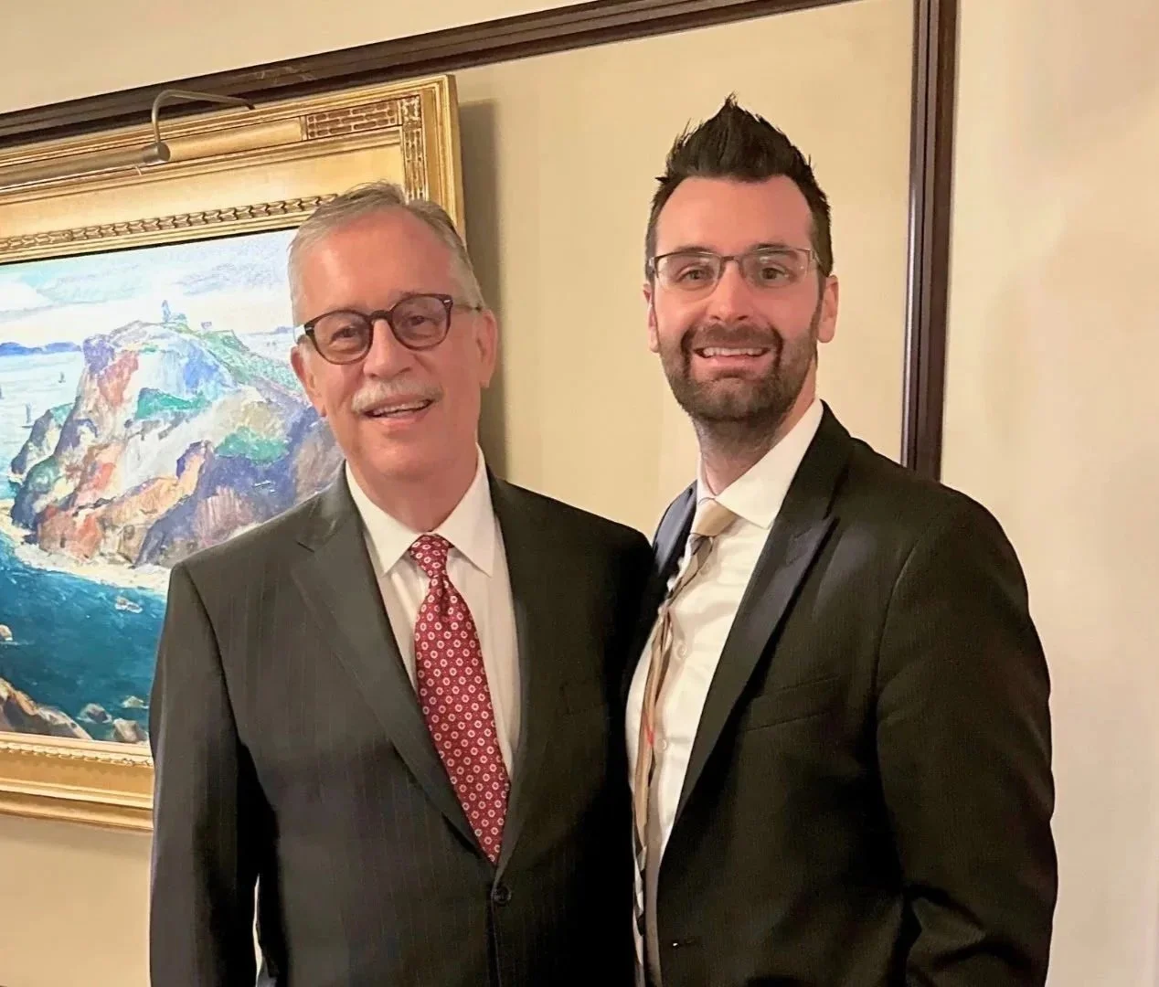 Two men in formal suits smiling indoors, with framed mountain landscape painting in the background.