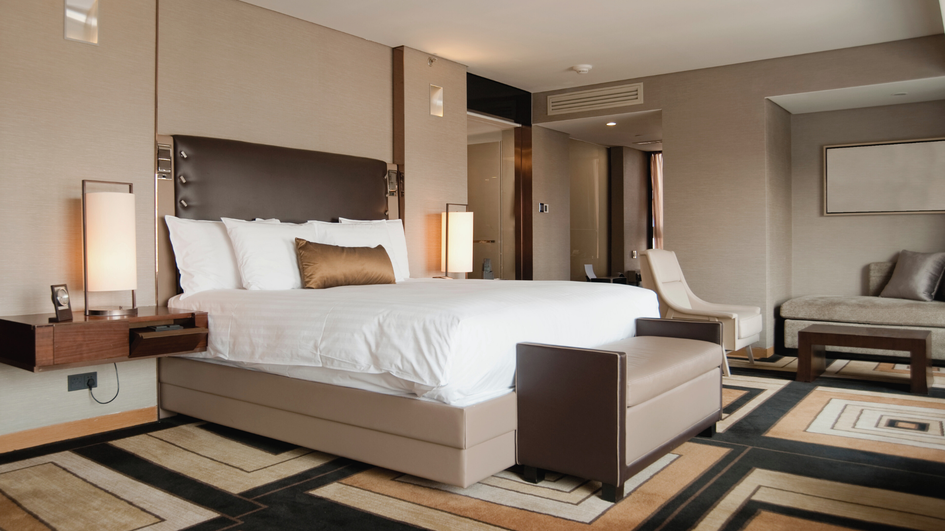 A modern hotel room featuring a large bed with white linens and a central brown pillow, bedside lamps, a beige chaise lounge, and a sofa with gray pillows, with neutral-colored walls and patterned carpet.